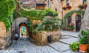 Un vicolo acciottolato in un borgo da visitare, con case in pietra ricoperte di edera e fiori. Un arco conduce a una vista panoramica.