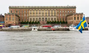 Stor byggnad vid vattnet, troligen kuninkaanlinna tukholma, med bussar och båtar. Svenska flaggan vajar.