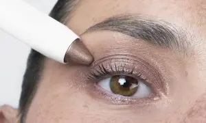 Application d'un fard à paupières bronze sur des yeux marrons. Un bâtonnet blanc dépose la couleur sur la paupière.