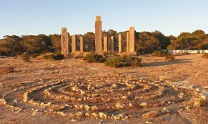 Steine bilden ein Labyrinth im Freien, umgeben von Pinien. Im Hintergrund ragen steinerne Säulen auf, die an Zeit und Raum auf Ibiza erinnern.