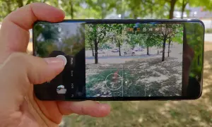 Dłoń trzyma smartfon z otwartą aplikacją do robienia zdjęć. Widok na drzewa i samochody.