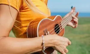 Kobieta w żółtej koszulce gra na ukulele na tle zielonej łąki i morza.