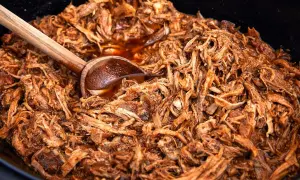 En stor gryta med saftig, långkokt pulled pork i en mörk sås. En träsked vilar i den läckra pulled pork grytan.