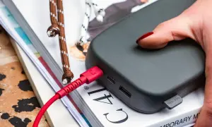 Ciemnoszary power bank 50000 mah na ile starcza? Podłączony czerwonym kablem, leży na stosie książek.