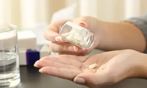 Kobieta wysypuje kapsułki z buteleczki na dłoń. Czy paracetamol jest przeciwzapalny? Obok stoi szklanka wody i pudełka leków.