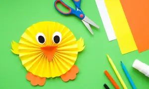 Un pollito de papel, ideal para manualidades fáciles para niños de 3 a 6 años. Tijeras, rotuladores y cartulinas de colores completan la escena.