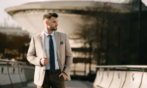 Elegancki mężczyzna w beżowej marynarce, na który guzik zapina się marynarkę, stoi na tle nowoczesnej architektury.