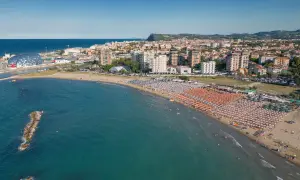 Vista aerea della Pesaro strand, con ombrelloni colorati, mare azzurro e la città sullo sfondo.
