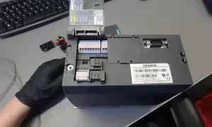 Ręka w czarnej rękawiczce trzyma sterowniki Siemens Sinamics G120C PN. Widać złącza, porty USB i etykietę z numerem seryjnym.