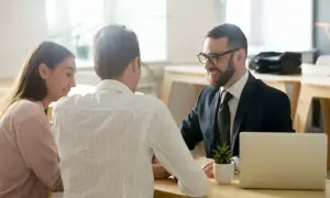 Coppia sorridente discute con un consulente di surroga mutuo consap, con laptop e pianta sul tavolo.