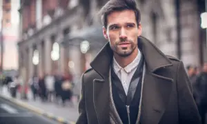Ein Mann mit Bart trägt eine elegante, olivgrüne Jacke zum Anzug. Er blickt nach rechts, während er durch eine belebte Stadt geht.