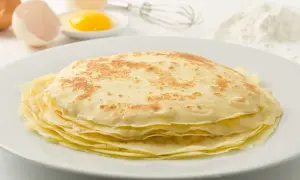 Ein Stapel goldbrauner Palatschinken auf einem Teller, daneben Eier, ein Schneebesen und Mehl. Perfekt für ein Palatschinken Rezept!