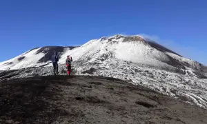 Dwie osoby stoją na zboczu wulkanu Etna pokrytego śniegiem. W tle widać dymiący krater.