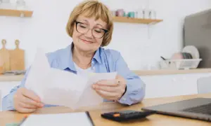 Uśmiechnięta starsza pani w okularach przegląda dokumenty, przygotowując wniosek o emeryturę. Na stole leży kalkulator i laptop.