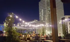 Abendstimmung auf einer Dachterrasse in Berlin. Lichterketten beleuchten Tische, an denen Gäste sitzen. Im Hintergrund ragen moderne Gebäude auf, die an den Spreegold Bikini erinnern.