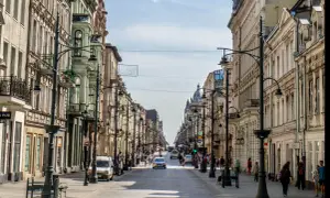 Belebte Straße in **Łódź, Polen**, gesäumt von historischen Gebäuden und Straßenlaternen. Autos und Fußgänger beleben die Szene.