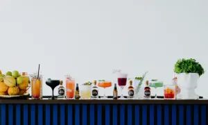 Rząd kolorowych drinków i butelek z alkoholami na barze, obok koszyka z owocami i białej rzeźby głowy.