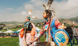 Zwei junge Menschen in traditioneller Kleidung mit Federschmuck und Trommeln. Sie nutzen die Gelegenheit, ihre Kultur zu präsentieren.