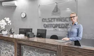 Recepcjonistka w okularach stoi przy ladzie w nowoczesnej klinice ortopedycznej.