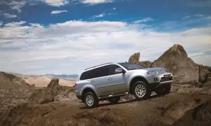 Srebrny Mitsubishi Pajero Sport pokonuje skalisty teren na tle pustynnego krajobrazu i błękitnego nieba z chmurami.