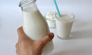 Domowy mleko kokosowe przepis. Butelka pełna białego napoju, obok szklanka ze słomką i miseczka z wiórkami kokosowymi.