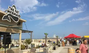 Strandpromenade in Den Haag mit dem Restaurant "The Fat Mermaid", Menschen, Sonnenschirmen und einem Riesenrad. Tolle Den Haag Unternehmungen!