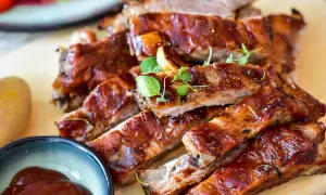 Saftige Rippchen, mariniert zum Grillen, glänzen in einer süßen BBQ-Sauce. Daneben eine Schale mit Sauce und eine Kartoffel.