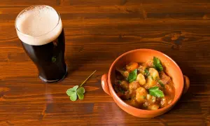 Un bicchiere di birra stout scura con schiuma cremosa accanto a una ciotola di stufato irlandese e un quadrifoglio su un tavolo di legno.