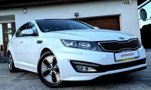 Biały kia sedan z czarnym dachem, zaparkowany przed domem z drewnianą bramą garażową.