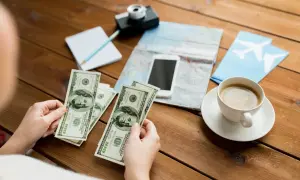 Ręce trzymające banknoty 100 USD, obok mapa, telefon, aparat, notatnik i kawa. Planowanie podróży, a niewypłacanie świadczenia urlopowego co to znaczy?