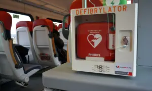Czerwony defibrylator AED w białej obudowie, zamontowany w pociągu, obok siedzeń.