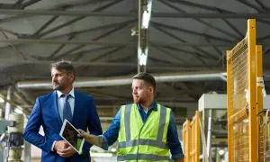 Mężczyzna w garniturze i pracownik w kamizelce odblaskowej omawiają stan bezpieczeństwa i higieny pracy w zakładzie, patrząc na tablet.