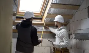 Dwóch budowlańców w kaskach mierzy otwór okna dachowego, planując montaż styropianu na poddasze.