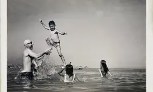 Un bambino salta in aria, sorretto da un uomo con occhiali da sole, mentre due bambine nuotano. Un momento di gioia estiva, un ricordo di Carlo Traini.
