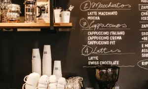 Tafel mit Kaffeebar Ideen: Macchiatos, Cappuccinos, Lattes und mehr. Stapel von Pappbechern und Espressomaschine im Vordergrund.