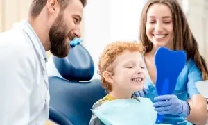 Uśmiechnięty chłopiec ogląda swoje zęby w lusterku w kształcie zęba, podczas gdy dentysta i asystentka patrzą z aprobatą. Czy fluoryzacja jest szkodliwa? Nie, to zabieg chroniący zęby.