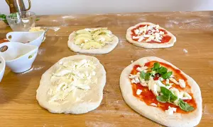 Vier rohe Pinsa-Teige auf einem Holztisch, teils belegt mit Tomatensoße, Mozzarella, Äpfeln und Basilikum.