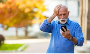 Anziano con mano sulla testa, guarda il telefono con espressione preoccupata, forse per la paura di parlare al telefono.