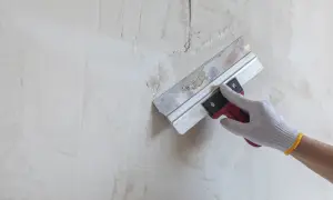 Ręka w rękawicy nakłada masę szpachlową na ścianę, wypełniając głębokie pęknięcia.