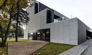 Nowoczesny budynek z metalicznym ryzalitem, przeszklonym wejściem i drewnianym tarasem porośniętym jesiennymi liśćmi.