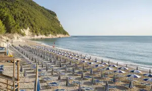 Filettata di ombrelloni e lettini sulla spiaggia di San Michele, con il mare azzurro e la collina verde sullo sfondo.