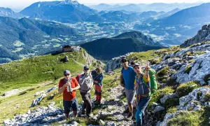 Wandergruppe auf dem Weg zum hocheck Watzmann. Im Hintergrund ein Alpenpanorama mit einem Dorf.