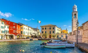 Colorful buildings line a harbor with boats, an Italian flag, and a bell tower under a clear blue sky.