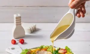 Ręka polewa sałatkę kremowym sosem. W tle stoją ozdobne solniczki i dwa pomidorki koktajlowe.