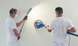 Deux ouvriers en combinaison blanche utilisent des ponceuses pour mur. L'un ponce le haut du mur, l'autre le centre.