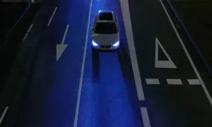 Un coche blanco con las luces encendidas, conduciendo de noche por una carretera oscura con marcas viales blancas y un reflejo azulado.