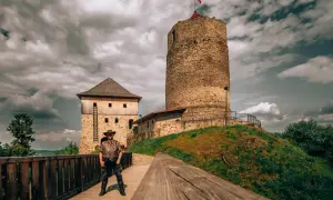 Zamek w Czchowie, z wieżą i budynkiem mieszkalnym, na tle burzowego nieba. Na drewnianym pomoście stoi mężczyzna w kapeluszu.