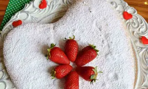 Torta panna e fragole velocissima a forma di cuore, cosparsa di zucchero a velo e decorata con fragole fresche e cuoricini rossi.