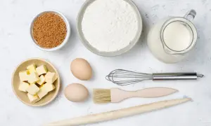 Ingredientes para hornear: harina, azúcar moreno, mantequilla en cubos, huevos, leche en jarra, batidor, brocha y rodillo de madera.