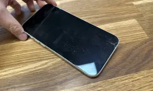 Dłoń trzyma telefon z pękniętym szkłem hartowanym. Ekran pokryty jest siecią drobnych pęknięć, szczególnie widocznych w prawym dolnym rogu.